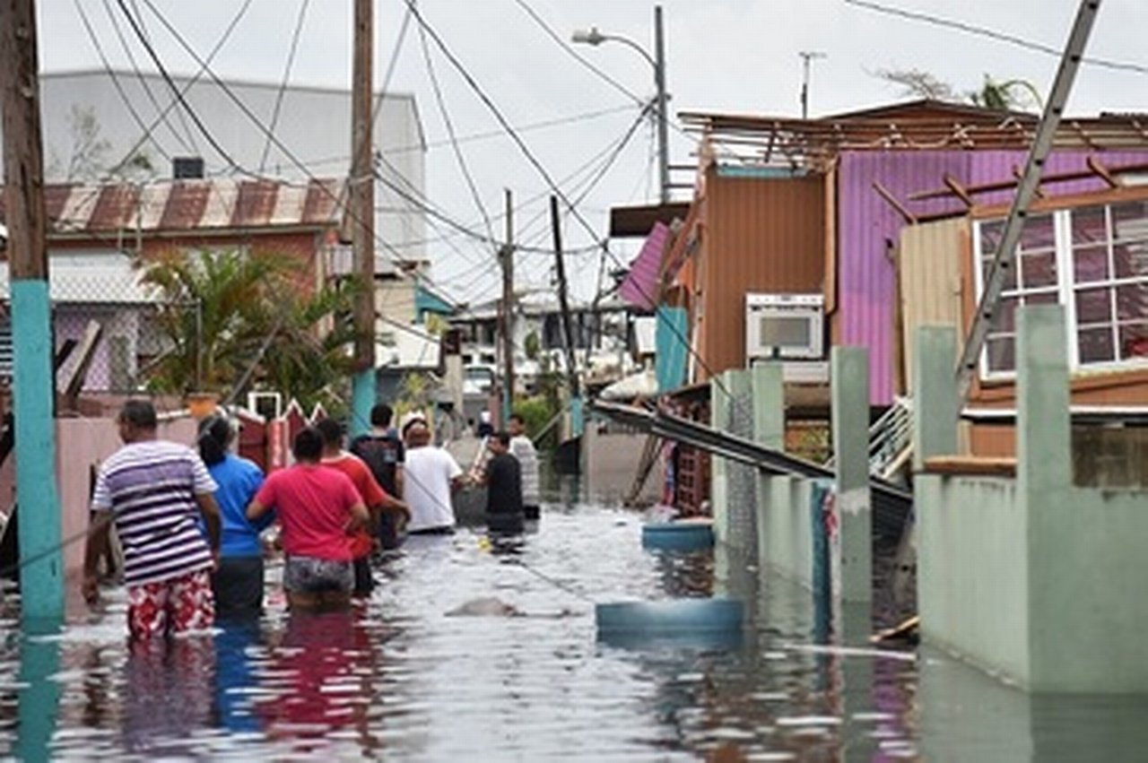 People walk in a flooded street next to damaged houses in Puerto Rico, where Hurricane Maria has created a humanitarian crisis. Photo credit: HECTOR RETAMAL/AFP/Getty Images